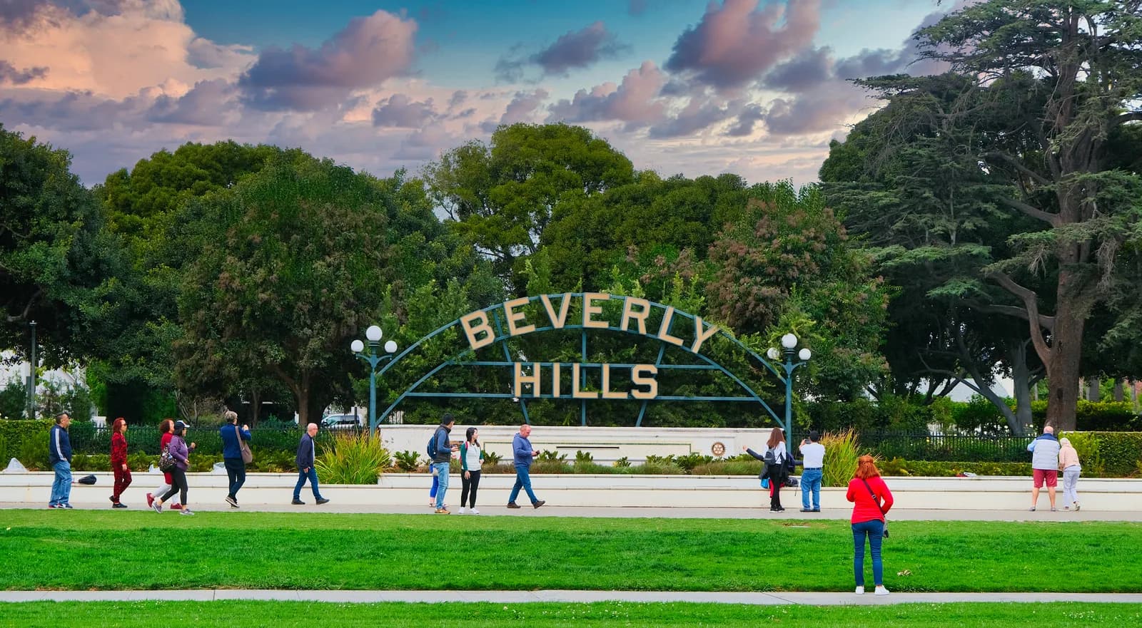 The wrought iron Beverly Hills park sign at dusk, with people walking on the lawn in front of it