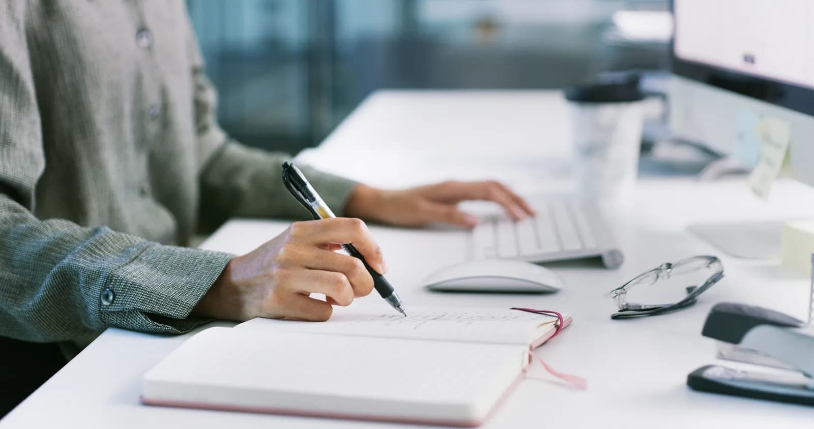 A hand writing in a notebook at a white desk, the other hand resting beside a keyboard and mouse