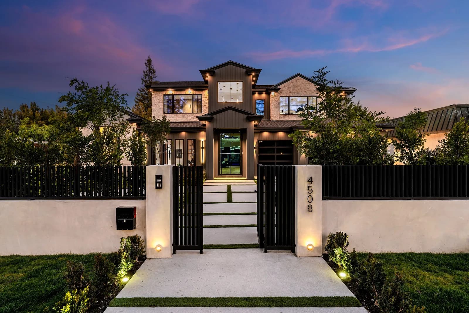 A modern estate lit at dusk behind a closed black gate, with the street number on the entrance pier