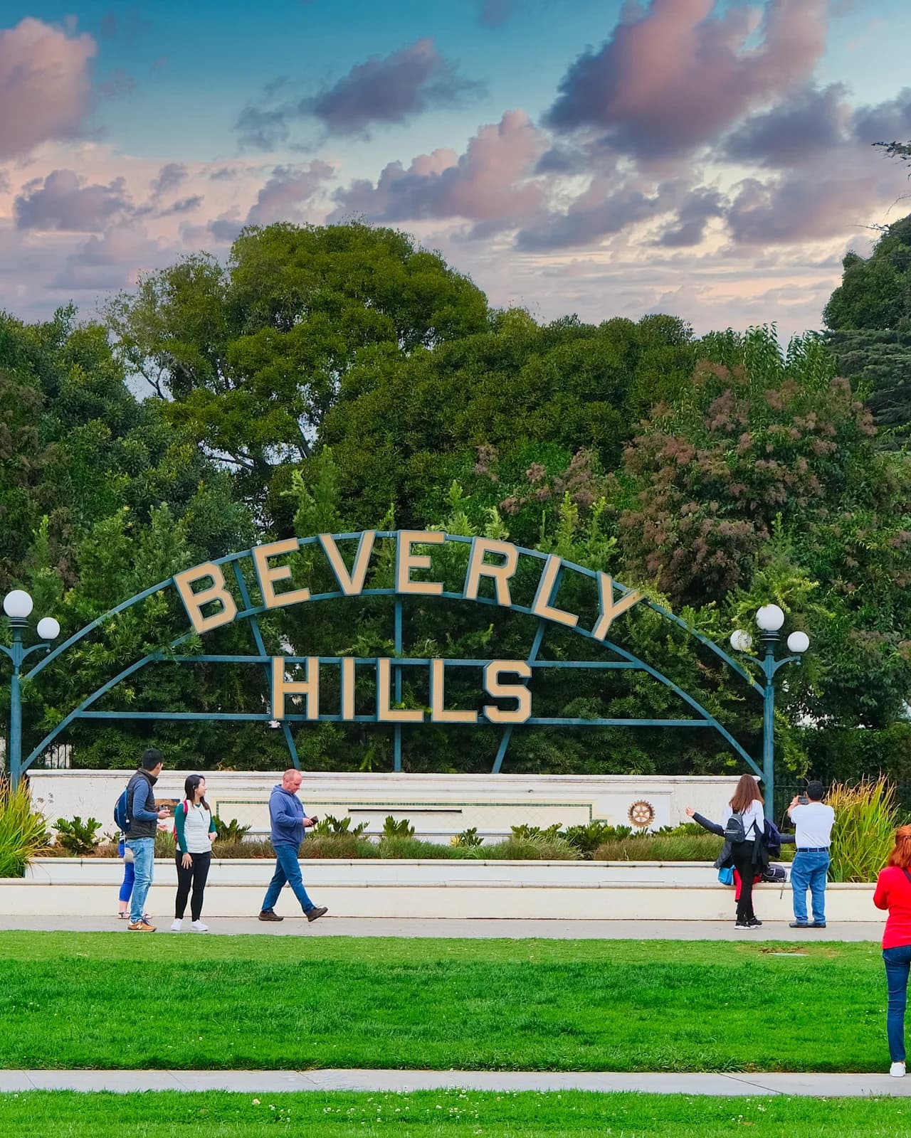 The wrought iron Beverly Hills park sign at dusk, with people walking on the lawn in front of it