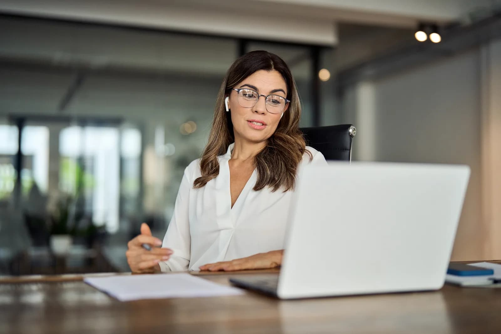 A woman in glasses working at a laptop across a desk in a bright office