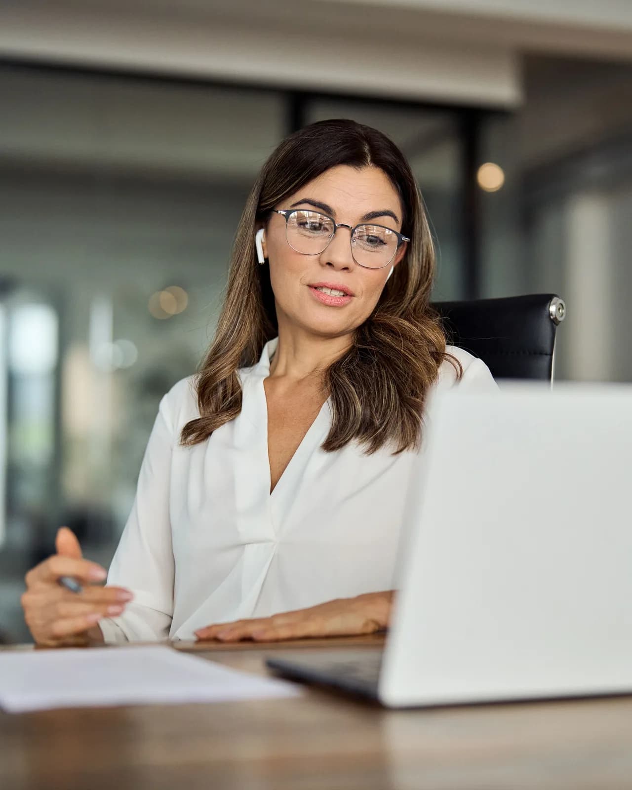 A woman in glasses working at a laptop across a desk in a bright office