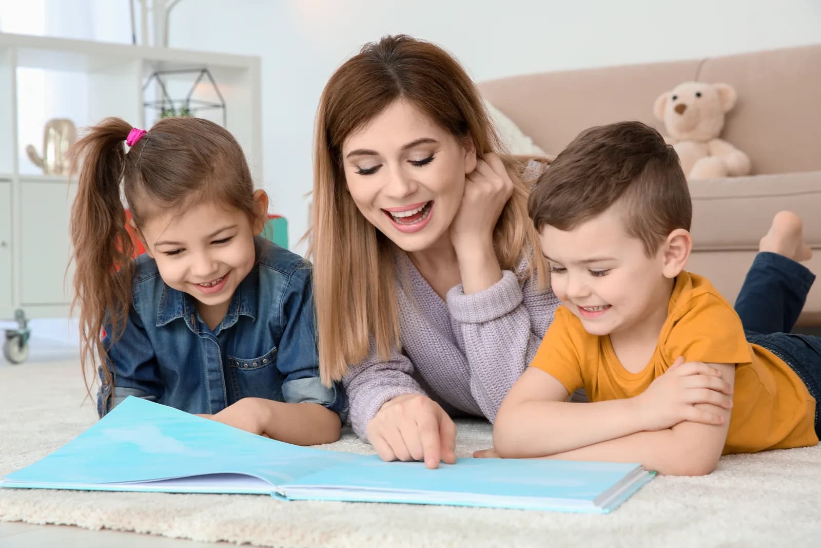 A woman, a young girl and a young boy lying on a rug, reading an open picture book together