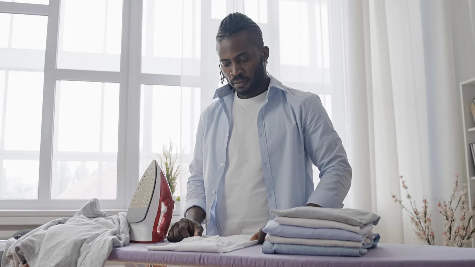 A man folding pressed shirts at an ironing board by a bright window, a red iron upright beside him