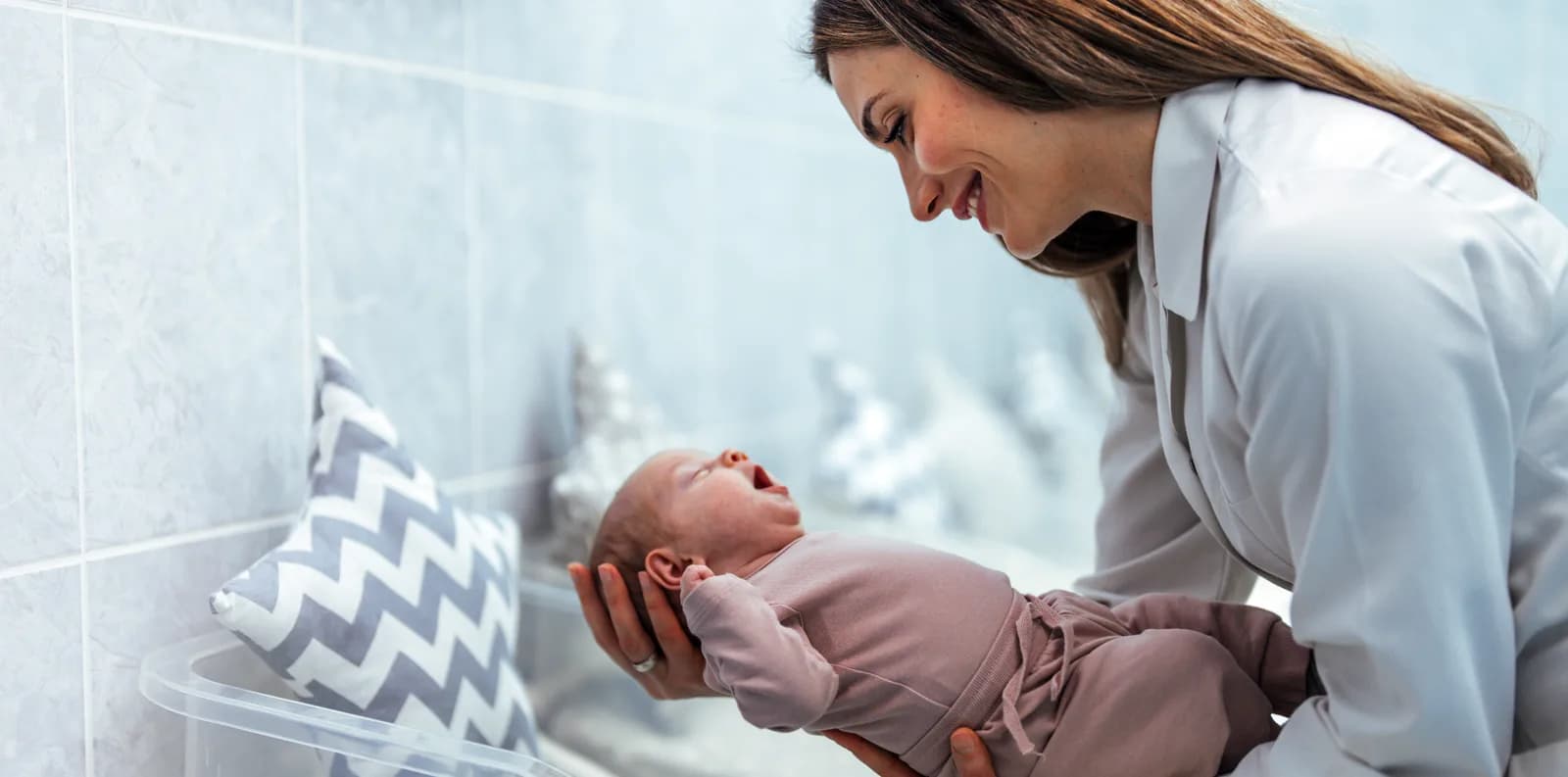 A woman leaning over a yawning newborn in a pink knit sleepsuit, held in her hands