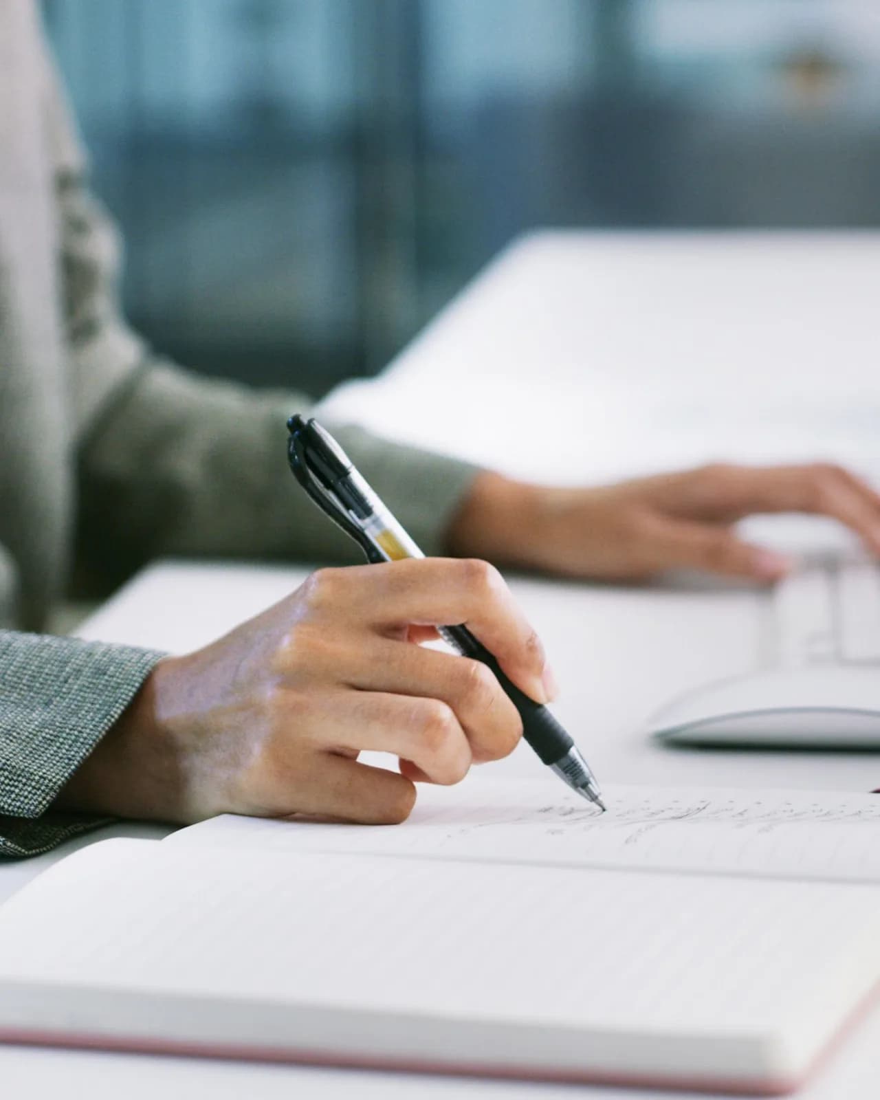 A hand writing in a notebook at a white desk, the other hand resting beside a keyboard and mouse
